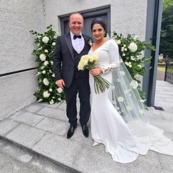 Real bride Emma stood with husband Brian by a cream floral decorated dark grey front door in Ronald Joyce 18202 Tai wedding dress. Emma has black hair tied up and red lips.
